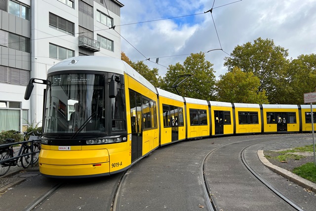 Ein moderner gelber Straßenbahnzug aus mindestens sieben Elementen fährt eine relativ enge Kurve, im Hintergrund ein mehrstöckiges Haus und Bäume.
